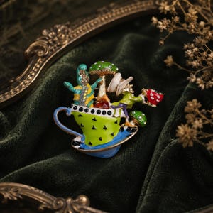 May include: A gold-tone metal brooch featuring a bright green teacup with black accents, containing a blue and yellow caterpillar, a tall green mushroom with white spots, and a figure in a hat holding a small red and white polka-dot teacup.