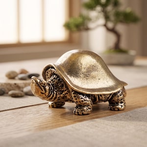 Puede incluir: Una pequeña figura de tortuga de metal en tono dorado con un caparazón liso y redondeado, además de cabeza y patas texturizadas y detalladas. La escultura presenta una postura realista con la cabeza extendida hacia adelante.