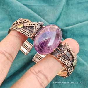 Puede incluir: Un brazalete de alambre de cobre envuelto con una gran piedra preciosa de amatista púrpura, ovalada y translúcida en el centro. El trabajo en metal incluye intrincados patrones de alambre enrollado y pequeños acentos circulares de latón.