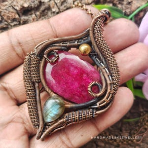 May include: A wire-wrapped pendant featuring a large, faceted, oval-shaped magenta gemstone and a smaller, teardrop-shaped iridescent gray stone. The frame is constructed from oxidized copper wire with intricate coils and a gold-toned bead. Text reads HANDMADEBKJEWELLERY.