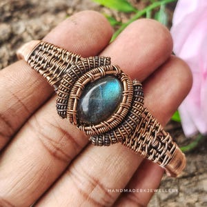 May include: A copper wire-wrapped cuff bracelet featuring an oval labradorite gemstone with blue iridescence at the center. The metalwork consists of intricate woven and coiled copper wire patterns. The text HANDMADEBKJEWELLERY is visible.