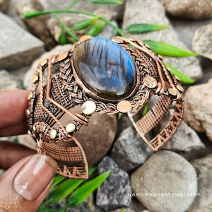 Puede incluir: Un brazalete de alambre de cobre trenzado con una gran piedra preciosa de labradorita ovalada con iridiscencia azul. El trabajo en metal incluye intrincados patrones tejidos, detalles de alambre trenzado y pequeños acentos circulares de cobre.