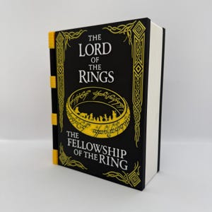 May include: A black book titled The Lord of the Rings The Fellowship of the Ring with yellow text and a yellow ring graphic on the cover. The spine features four yellow horizontal accents, and the page edges are white, creating a distinct contrast.