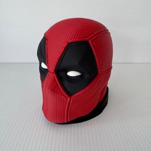 May include: A red and black full-head mask featuring a textured surface pattern. The mask has large, angular black eye patches with white oval-shaped eye openings, mimicking the appearance of the character Deadpool from comic book media.