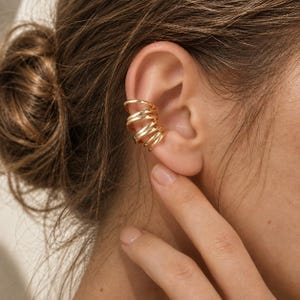 Puede incluir: Un ear cuff de tono dorado con un diseño en espiral de varias capas que se envuelve alrededor del cartílago de la oreja. La pieza consta de varias bandas lisas y pulidas que crean una apariencia apilada, ofreciendo un aspecto enrollado.