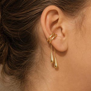May include: A gold-toned ear cuff featuring two smooth, parallel bands that wrap around the cartilage. Two delicate gold chains hang from the bands, each ending in a polished, teardrop-shaped pendant that dangles freely below the earlobe.