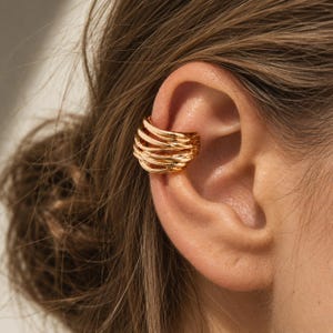 Op de afbeelding: Een goudkleurige ear cuff met een meerlaags, omhullend ontwerp bestaande uit verschillende gladde, met elkaar verbonden banden die langs het kraakbeen van het oor krullen. Het metalen oppervlak heeft een gepolijste afwerking die licht reflecteert.
