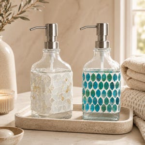 May include: Two square glass soap dispensers with silver-toned metal pumps. One dispenser features a white and gold mosaic tile pattern, while the other displays a blue and green leaf-shaped mosaic tile design, both resting on a rectangular tray.