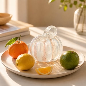 May include: A clear glass pumpkin sculpture with a curled stem and subtle white and orange speckled vertical stripes, positioned on a round plate alongside a whole orange with a green leaf, a yellow lemon, a green lime, and a slice of orange.