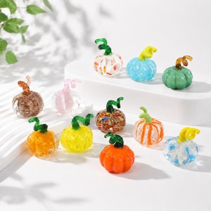 Puede incluir: Una colección de once pequeñas calabazas de vidrio soplado a mano en varios colores y patrones, incluyendo diseños sólidos en naranja, amarillo, azul claro y verde, así como modelos moteados, rayados y transparentes, cada uno con un tallo verde.