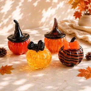 Peut inclure: Quatre figurines de citrouilles en verre. Une est orange avec un chapeau de sorcière noir, une est orange avec une tige marron foncé, une est jaune avec un nœud noir, et une est rayée noir et orange avec un nœud orange. Forme arrondie côtelée.
