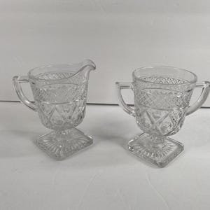 Op de afbeelding: Twee heldere glazen serveerstukken. Eén is een roomkannetje met een tuit en handvat, de andere is een suikerpot met twee handvatten. Beide hebben een ruitpatroon en staan op vierkante voetstukken. Ideaal om te serveren.