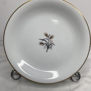 Könnte beinhalten: Weißer Keramikteller mit goldenem Rand und einem Weizenähren-Design in der Mitte. Der Teller wird auf einem Metallständer präsentiert. Ein klassisches Design zum Servieren von Speisen.