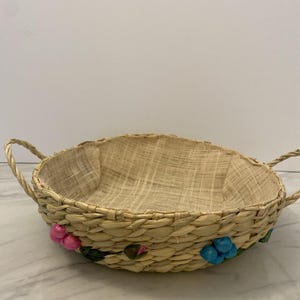 May include: Oval-shaped woven basket with two handles. The basket is made of natural-colored straw and features embroidered pink and blue floral accents along the rim. The interior is lined with a lighter-colored fabric.