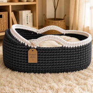Puede incluir: Una cesta ovalada de ganchillo en color gris oscuro con un borde blanco grueso y asas integradas. Una etiqueta de papel rectangular beige está atada al asa con cordel, con el texto "Handmade with love" y un pequeño corazón debajo.