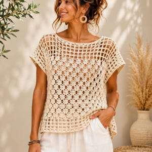 Puede incluir: Un top de ganchillo beige con un amplio escote barco, mangas cortas y un patrón cuadrado repetitivo en todo el cuerpo. La prenda tiene una construcción de punto abierto con un borde tipo rejilla en el dobladillo y las mangas, combinado con pantalones blancos.