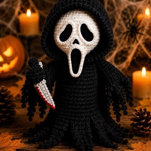 May include: A crocheted figure wearing a black hooded robe and a white mask with large black eye and mouth openings. The figure holds a small gray knife with a red-tipped blade in its right hand. The robe features a textured, fringed hem.