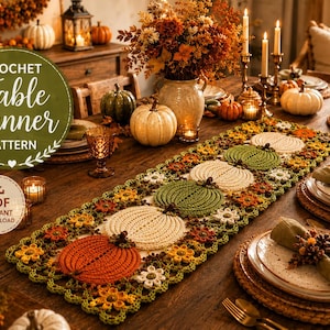 Puede incluir: Un camino de mesa de ganchillo con un patrón repetitivo de calabazas en naranja, crema y verde, decorado con pequeñas flores y hojas. El camino se muestra sobre una mesa de comedor de madera con platos y servilletas.