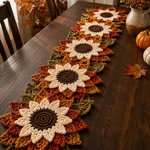 May include: A long, rectangular crochet table runner featuring a repeating pattern of sunflowers with dark brown centers and cream-colored petals, surrounded by layered leaves in shades of burnt orange, mustard yellow, and olive green.