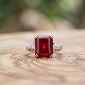 Peut inclure: Une bague argentée ornée d'une grande pierre rouge rectangulaire taille émeraude au centre. La bague est flanquée de deux pierres blanches transparentes plus petites, de taille baguette effilée, fixées par des griffes en métal.