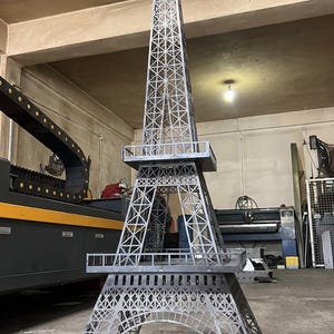 May include: A large, grey metal sculpture of the Eiffel Tower featuring a detailed lattice framework. The structure is composed of multiple levels with open platforms and a pointed spire at the top, showcasing industrial craftsmanship and metal construction.