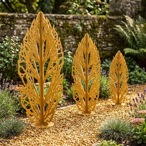 May include: Three mustard yellow metal garden sculptures shaped like stylized leaves with intricate cut-out patterns. Each sculpture stands on a circular base, arranged in a row from largest to smallest, featuring a central vertical spine.