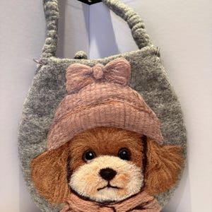 Può includere: Una borsa in feltro grigio con un ritratto in feltro ad ago di un cucciolo marrone e bianco che indossa un cappello rosa polvere con un fiocco e una sciarpa abbinata. La borsa ha una forma arrotondata e un manico in feltro grigio con anelli.