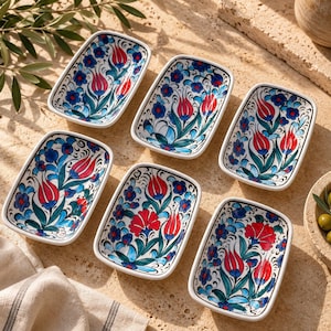 May include: A set of six rectangular ceramic dishes featuring a hand-painted floral pattern with vibrant red tulips and blue blossoms on a white background. Each small bowl has a smooth, glossy finish and rounded corners, arranged in a cluster.