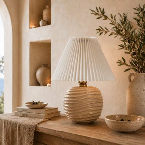 May include: A table lamp with a round, beige ceramic base featuring horizontal wavy ridges and a white, pleated, conical fabric lampshade. The lamp has a small, metallic gold-toned accent piece connecting the base to the shade.