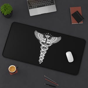 May include: A rectangular black desk mat featuring a white graphic illustration of a caduceus symbol. The design depicts a central staff entwined by two snakes, topped with a pair of wings and a small crown, surrounded by celestial stars and moons.