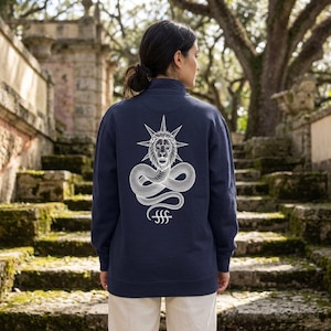 May include: A dark navy blue long-sleeve quarter-zip sweatshirt featuring a white graphic print on the back. The design displays a lion head centred above a coiled snake, topped with a starburst, and a small symbol consisting of three vertical lines.