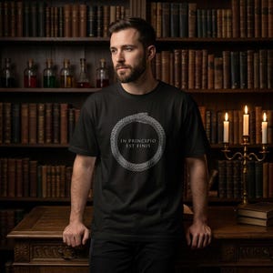 Puede incluir: Una camiseta negra de manga corta con un gráfico blanco de una serpiente Ouroboros formando un círculo. Dentro del círculo, el texto dice IN PRINCIPIO EST FINIS. El diseño está centrado en el pecho de la prenda, que tiene cuello redondo.