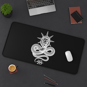 May include: A rectangular black desk mat featuring a white graphic print of a lion head with a starburst halo, attached to a coiled snake body, with a unique symbol underneath. The design is centered on the mat, which has rounded corners.