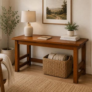 May include: A rectangular wooden console table with four sturdy legs and a smooth brown finish. The table surface holds a ceramic table lamp with a beige shade, a closed notebook with a pen, and two stacked books topped by a small potted plant.