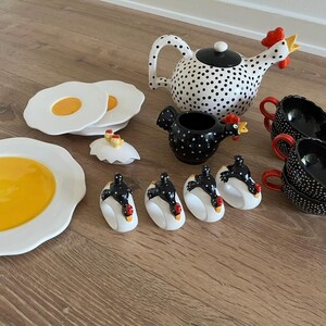 May include: A ceramic tea set featuring a white teapot with black polka dots and a rooster-shaped spout, a black creamer with white dots, four black cups with red handles, four egg-shaped napkin rings, and plates shaped like fried eggs.