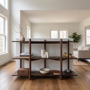 Peut inclure: Une étagère à trois niveaux en bois brun foncé avec des montants verticaux. Les étagères contiennent une pile de livres, un vase en céramique blanche, une horloge dorée, une petite plante succulente, une boîte en bois, un bol et un appareil photo.