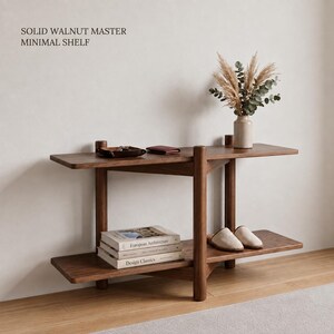 Könnte beinhalten: Ein Regal aus massivem Walnussholz mit zwei horizontalen Ebenen und vertikalen Stützen. Der Text SOLID WALNUT MASTER MINIMAL SHELF ist sichtbar. Das Regal enthält eine beige Vase mit getrockneten Pflanzen, ein kleines Tablett, eine Geldbörse, drei Bücher und zwei Hausschuhe.