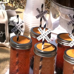 May include: Four glass jars with orange liquid and black and white striped straws. Each straw has a black and white sign that says "Rail Crossing Road".