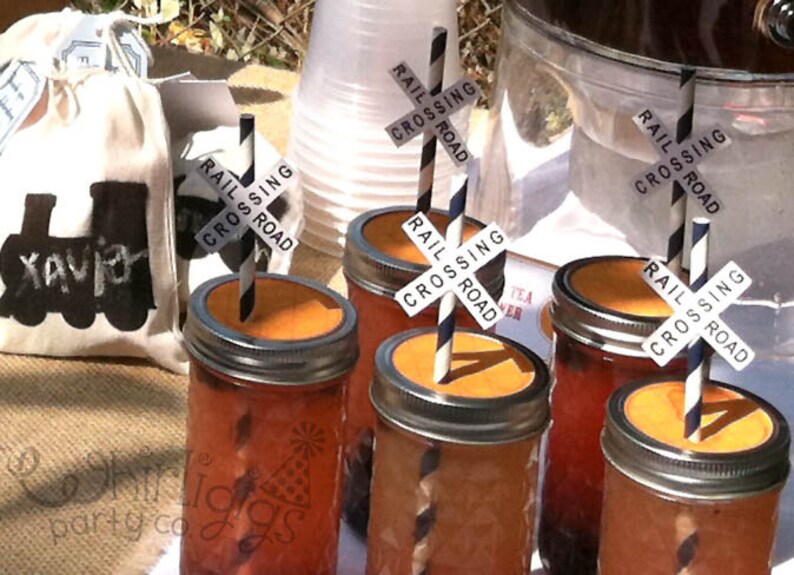 May include: Four glass jars with orange liquid and black and white striped straws. Each straw has a black and white sign that says "Rail Crossing Road".