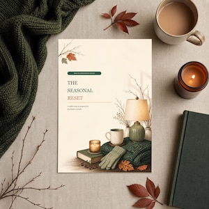 May include: A vertical rectangular print featuring the text THE SEASONAL RESET, a table lamp with a green base, a white mug, a lit candle on a book, and green knit gloves. The design includes autumn leaves and branches on a cream background.