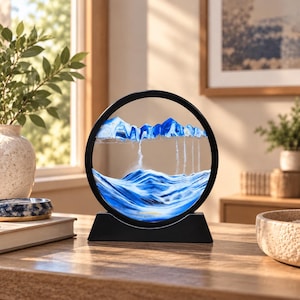 May include: A circular black frame containing a dynamic sand art display with layers of blue and white sand. The sand forms mountain-like peaks at the top and flowing, wave-like patterns at the bottom, encased within two clear glass panels.