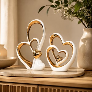 Puede incluir: Dos esculturas decorativas de corazones hechas de cerámica blanca con detalles en tono dorado pulido. La escultura más alta presenta un bucle alargado con un corazón pequeño, mientras que la más corta muestra formas de corazón anidadas con un corazón dorado central.
