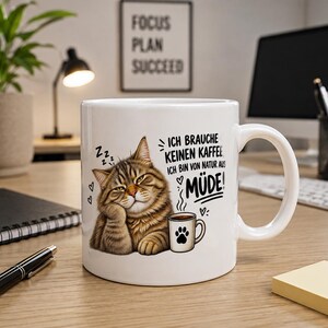 Könnte beinhalten: Ein weißer Keramikbecher mit einer Grafik einer müden, braunen getigerten Katze, die ihren Kopf auf die Pfote stützt. Neben der Katze ist eine kleine Tasse mit Pfotenabdruck abgebildet. Der Text lautet: ICH BRAUCHE KEINEN KAFFEE ICH BIN VON NATUR AUS MÜDE!