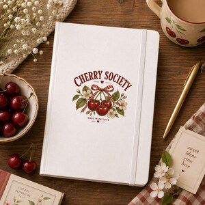 Pode incluir: Um caderno branco com fecho elástico apresentando um gráfico central de cerejas vermelhas, folhas verdes e um laço xadrez vermelho com o texto CHERRY SOCIETY e MADE WITH LOVE impresso em tinta vermelha na capa do caderno.