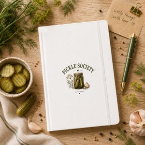 Könnte beinhalten: Ein weißes Notizbuch mit Gummibandverschluss, das mit einer Grafik eines Gurkenglases, Dill und Knoblauch sowie dem Text "PICKLE SOCIETY" und "MADE WITH LOVE" auf dem Einband versehen ist. Das Notizbuch ist von frischem Dill, Knoblauch und einem Stift umgeben.