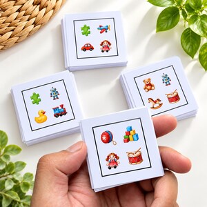 May include: A set of square white cards featuring colourful illustrations of various toys including a puzzle piece, aeroplane, car, doll, robot, rubber duck, train, teddy bear, rocking horse, drum, yo-yo, and building blocks, each enclosed in a black border.