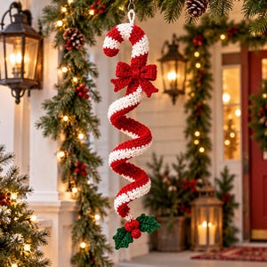 May include: A crocheted candy cane ornament featuring a red and white spiral design, accented with a red crocheted bow at the top and a cluster of three red berries with two green holly leaves attached to the bottom end of the spiral.