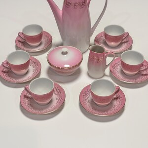 Könnte beinhalten: Ein Porzellan-Teeservice bestehend aus einer hohen Kaffeekanne, einer Zuckerdose mit Deckel, einem Milchkännchen und sechs Tassen mit passenden Untertassen. Jedes Teil hat eine weiße Basis mit rosa Farbverlauf, Goldrand und goldenem Rankenmuster.