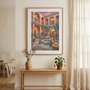 May include: A framed wall art print depicting a Moroccan-style courtyard with intricate blue and orange tile patterns, a central stone fountain, arched walkways, potted palm trees, and warm glowing lanterns under a soft sunset sky.