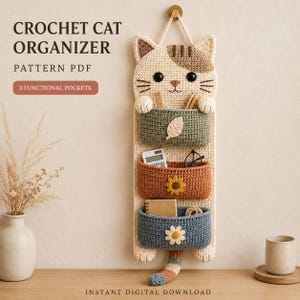 May include: A wall-hanging crochet cat organizer featuring three functional pockets in sage green, terracotta, and slate blue. The cat face is cream-colored with brown patches, black eyes, and whiskers. Text reads: CROCHET CAT ORGANIZER PATTERN PDF.